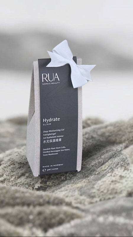 A small gray and white RUA skincare pouch labeled 'Hydrate' with a white ribbon bow, standing on textured gray rocks against a soft, blurred beach or water background.