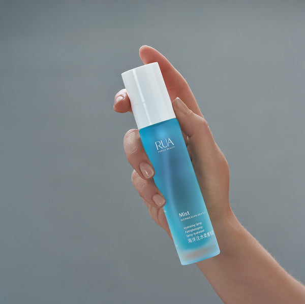 Hand holding a RUA Nordic Beauty - Mist Norwegian Sea Silk bottle with white reusable spray top against a gray background