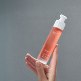 Hand holding a RUA Nordic Beauty  Remove Cleanser bottle with white reusable pump against a gray background